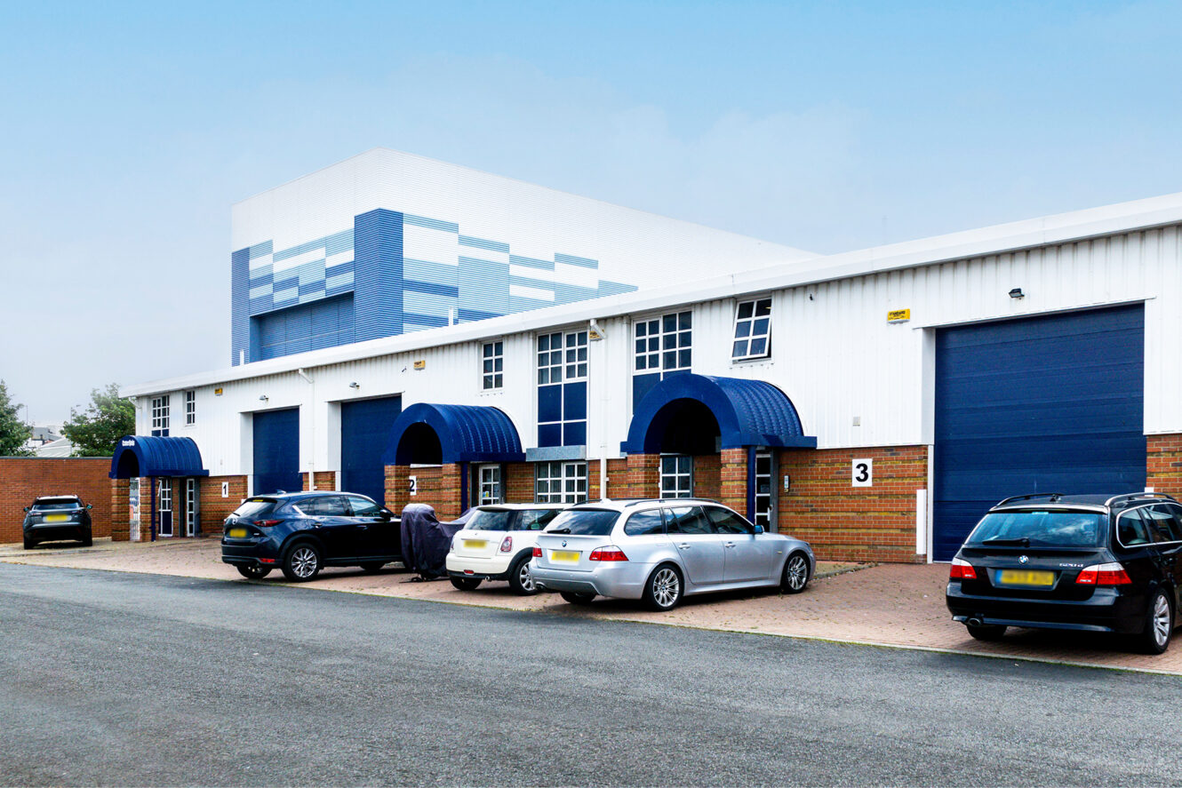 A row of commercial warehouse units with blue doors and awnings, several parked cars in front, and a clear sky overhead.