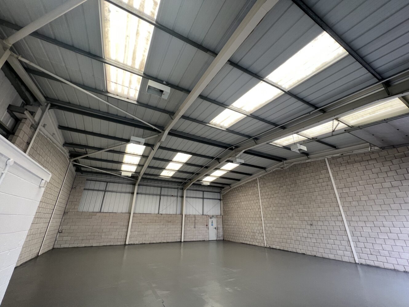Empty industrial warehouse with concrete block walls, gray painted floor, and a metal roof with several skylights allowing natural light inside.