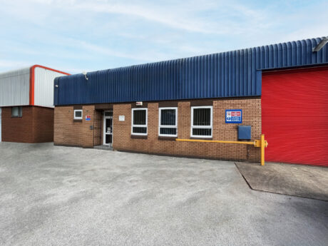 Single-story industrial building with brown brick walls, blue corrugated roof, three windows, an entrance door, and a large red roller shutter on the right.