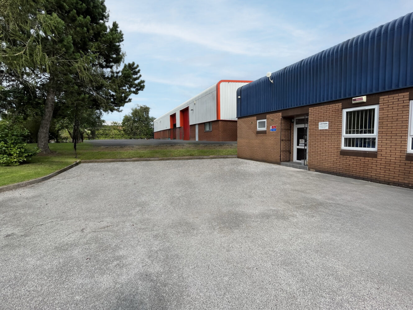 Single-story industrial buildings with brick and metal exteriors, adjacent grassy area, and an open doorway under a blue awning.