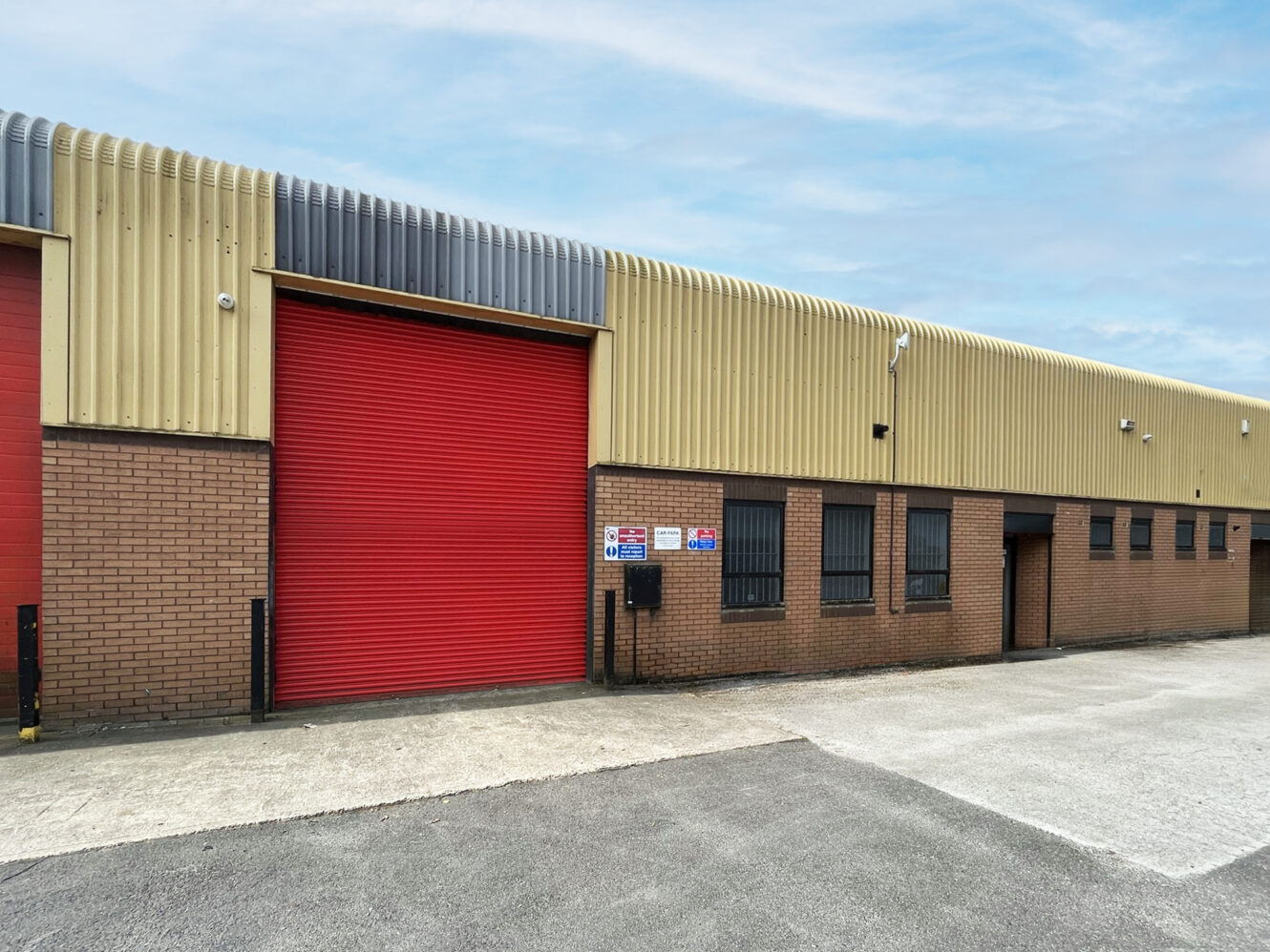A single-story industrial building with a corrugated metal roof, brick walls, and a large red roller shutter door, adjacent to a paved area.