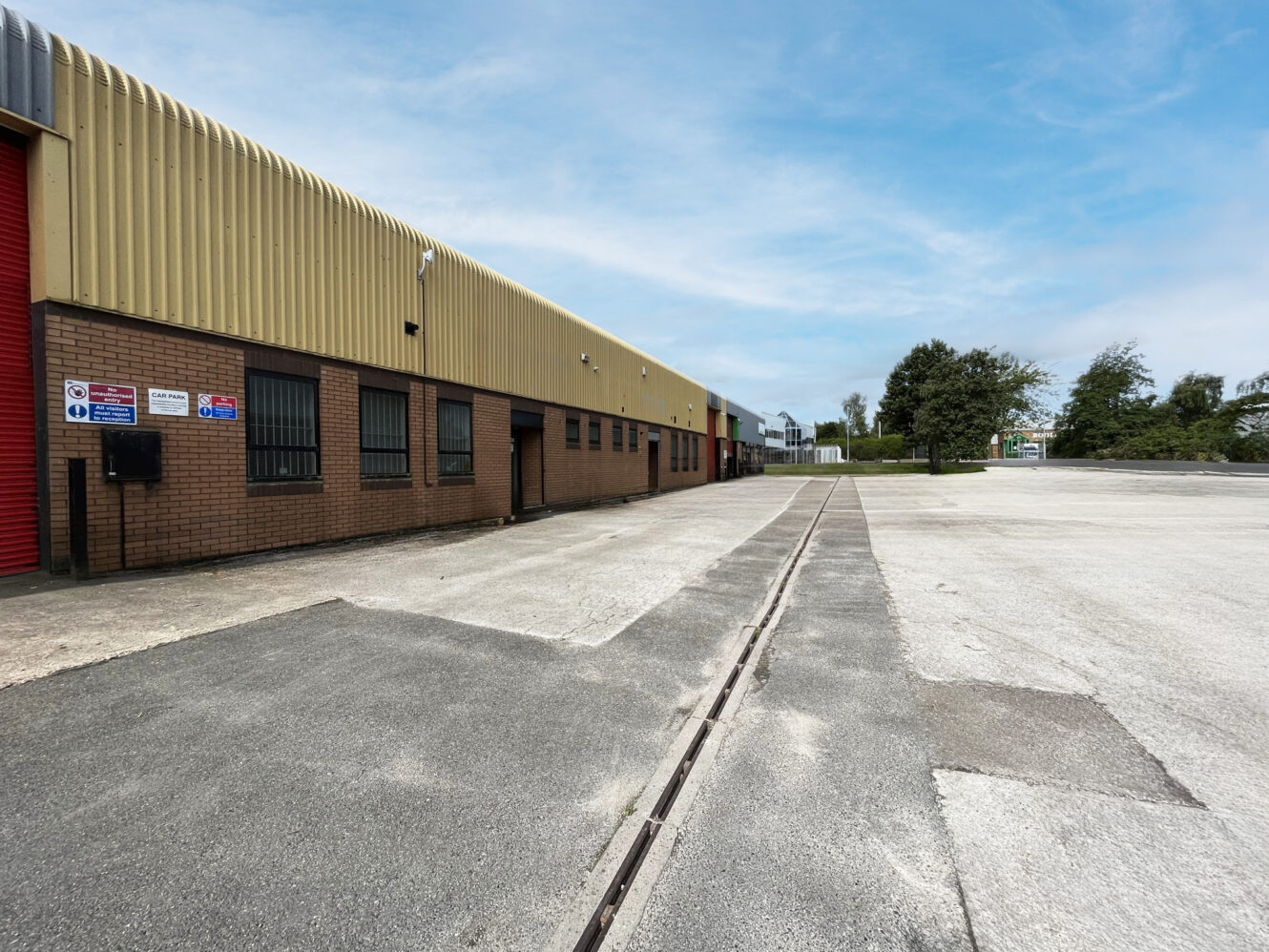 A row of industrial warehouse units with yellow and brown exteriors, red doors, security signs, and an empty paved lot under a blue sky.