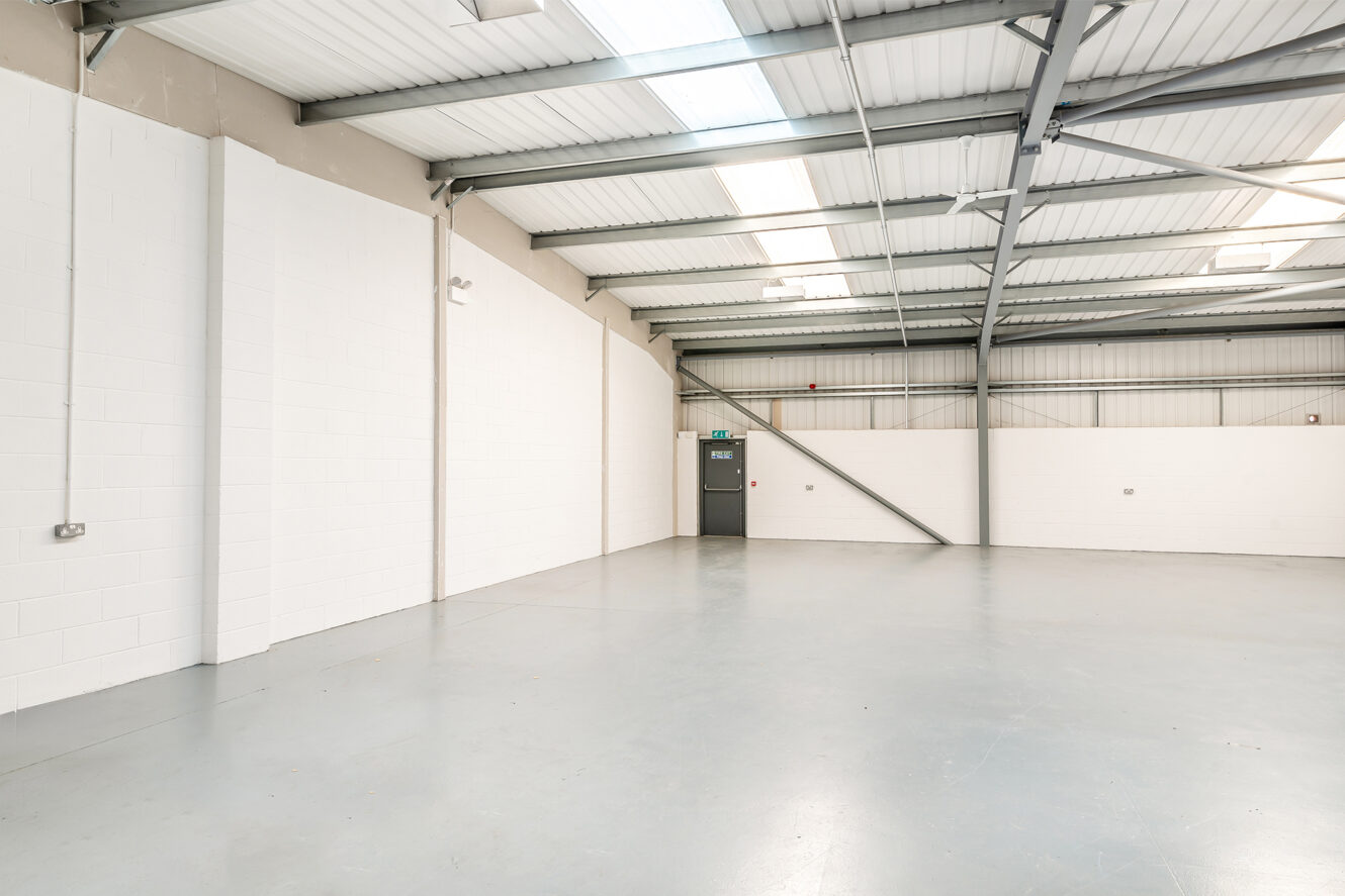 Spacious, empty industrial warehouse with white walls, grey steel beams, concrete floor, and a grey exit door under fluorescent lighting.