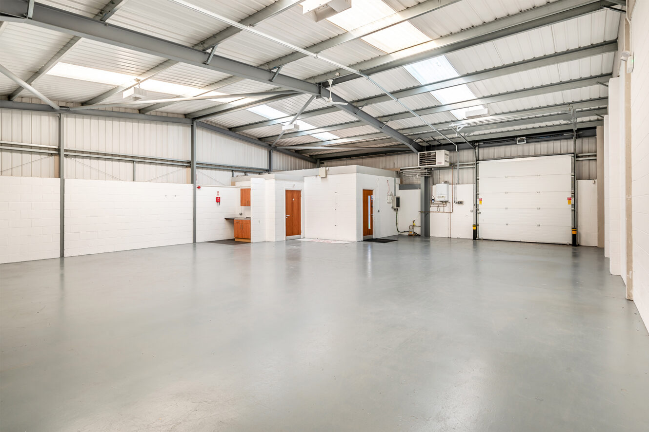 Interior of an empty industrial warehouse with a concrete floor, white brick walls, metal ceiling, office units, and a large roller door.