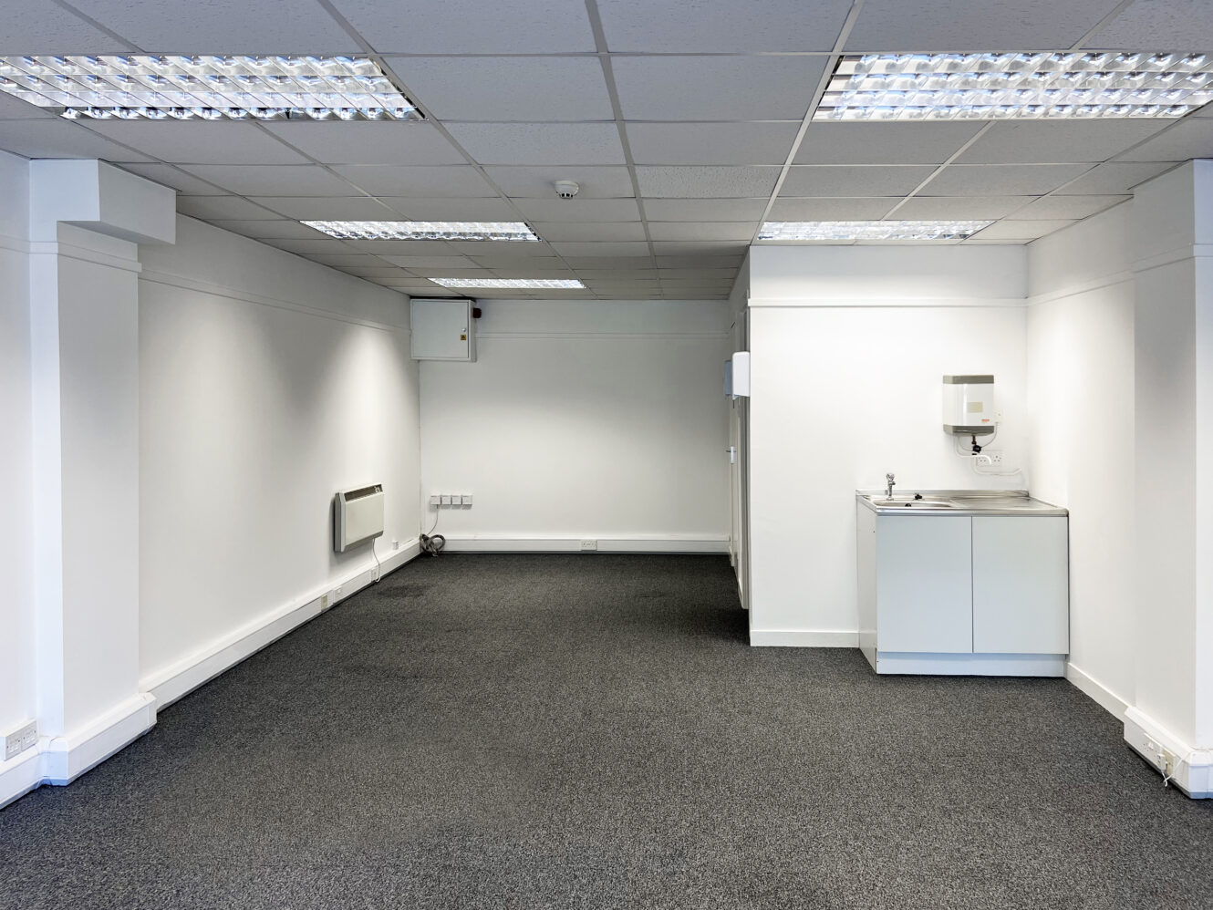 An empty office room with white walls, grey carpet, overhead fluorescent lights, a sink unit, wall heater, and electrical outlets.