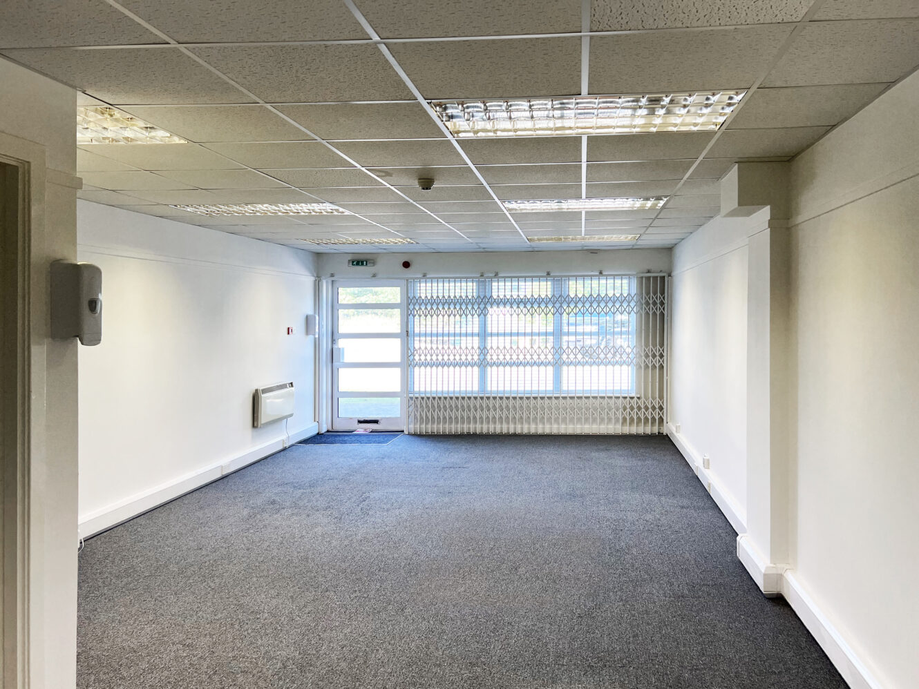 Empty office room with white walls, gray carpet, fluorescent ceiling lights, and a window with metal security bars at the far end.