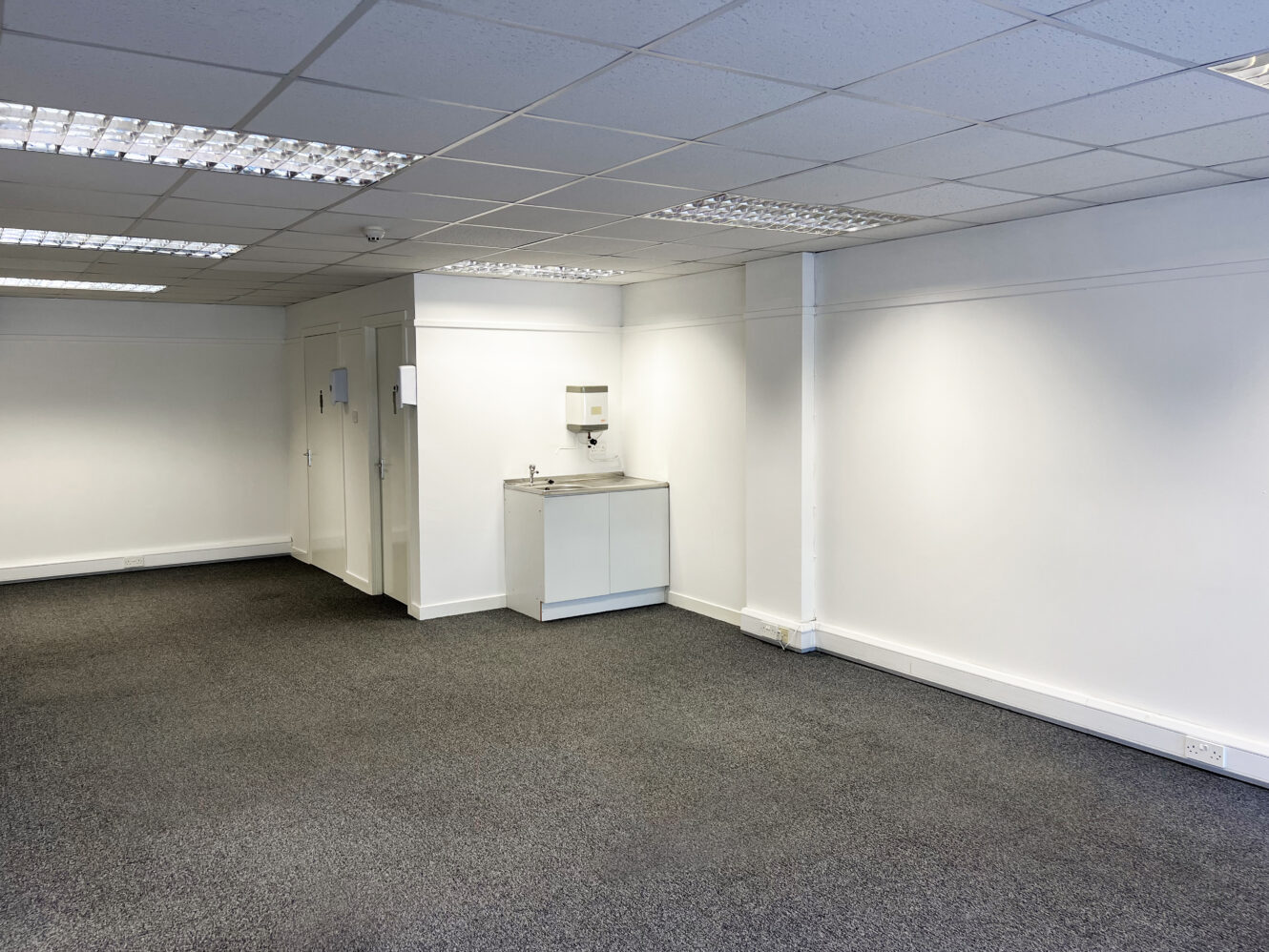 Empty office space with white walls, gray carpet, fluorescent ceiling lights, and a small kitchenette with a sink and wall-mounted soap dispenser in the back corner.