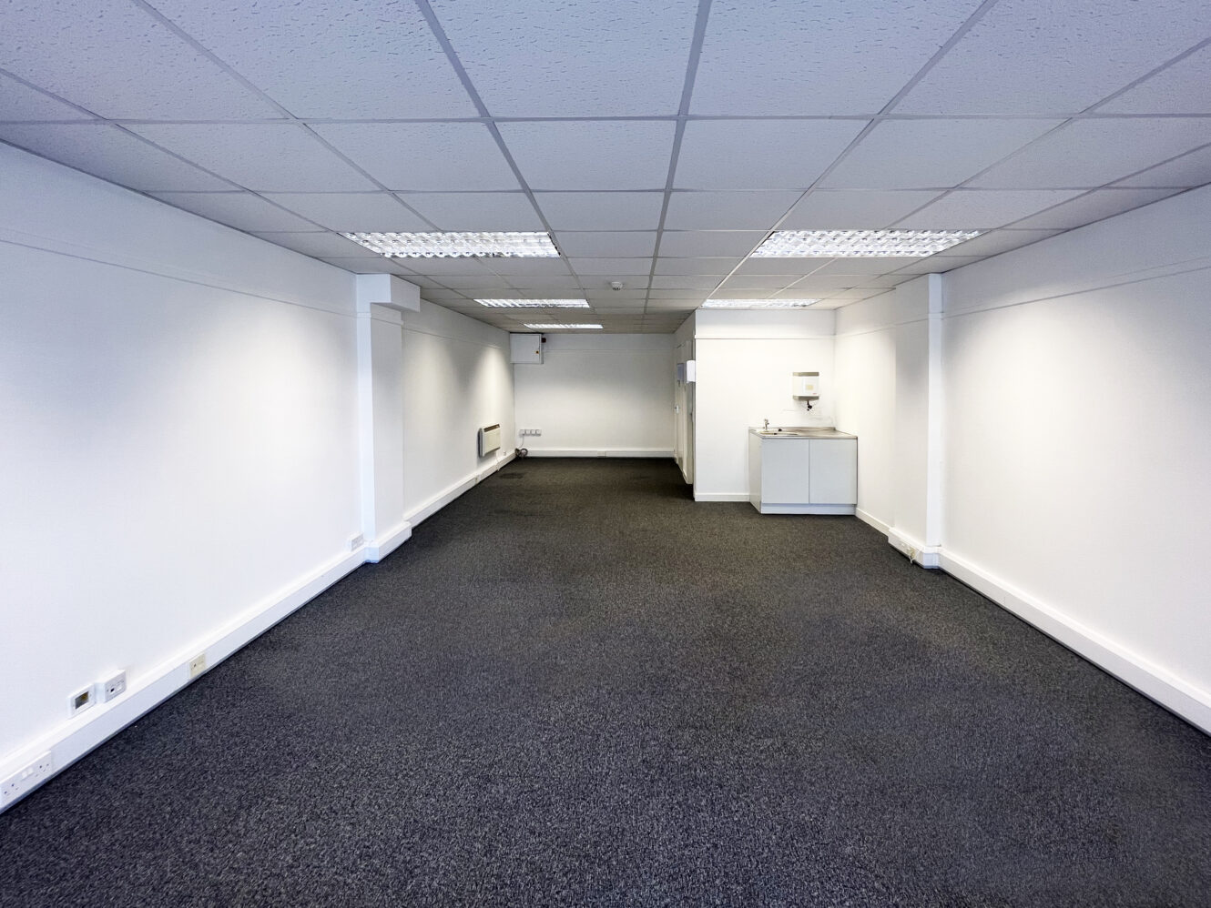 Empty office space with white walls, grey carpet flooring, suspended ceiling tiles, fluorescent lighting, and a small kitchenette area at the back.