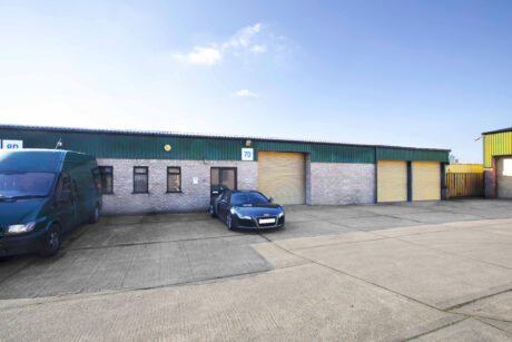 A black car and a green van are parked outside a row of industrial warehouse units with yellow roller doors and brick walls under a blue sky.