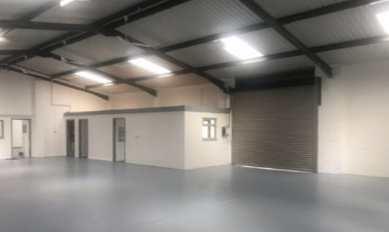 Interior of an empty industrial warehouse with a small office structure, a metal roller shutter door, and exposed beams on the ceiling.