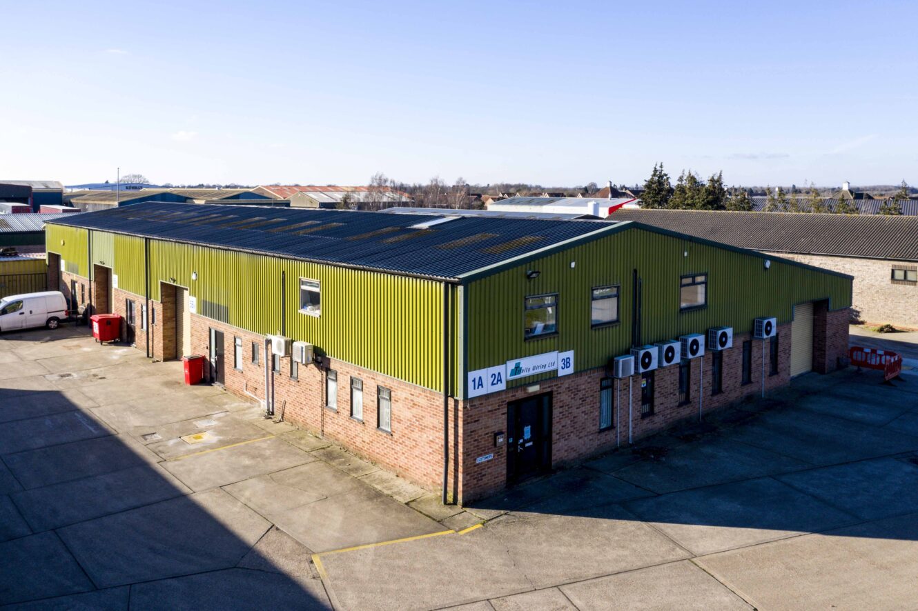 A single-story industrial warehouse with a green metal roof and brick walls, featuring multiple units and exterior air conditioning units, located in an industrial area.