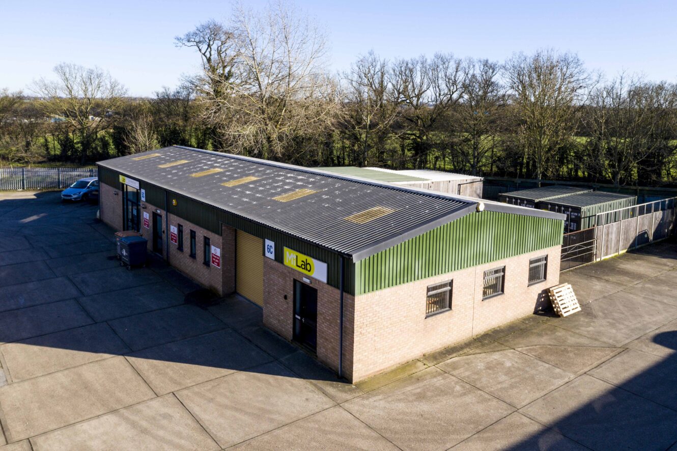 Aerial view of a single-story industrial building with multiple garage doors and a fenced area, surrounded by trees and an empty concrete lot.