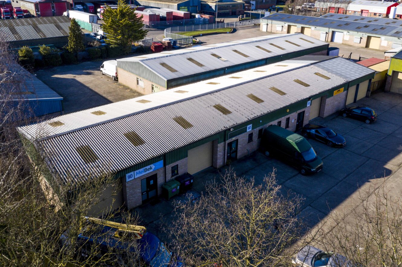 Aerial view of industrial units with corrugated metal roofs, parked vehicles, and signs on the front of the buildings.