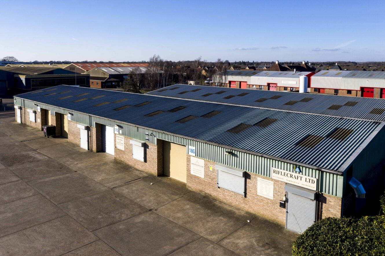 A row of single-story industrial warehouse units with roller shutter doors and corrugated metal roofs in a commercial area.