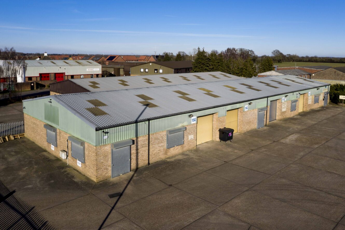 A row of industrial warehouse units with corrugated metal roofing and roller shutter doors, situated in a paved lot on a clear day.