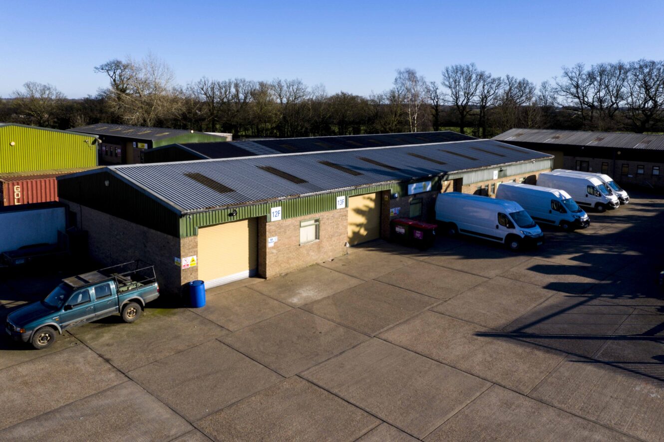 A row of industrial warehouse units with vehicles, including vans and a pickup truck, parked outside on a concrete lot under a clear blue sky.