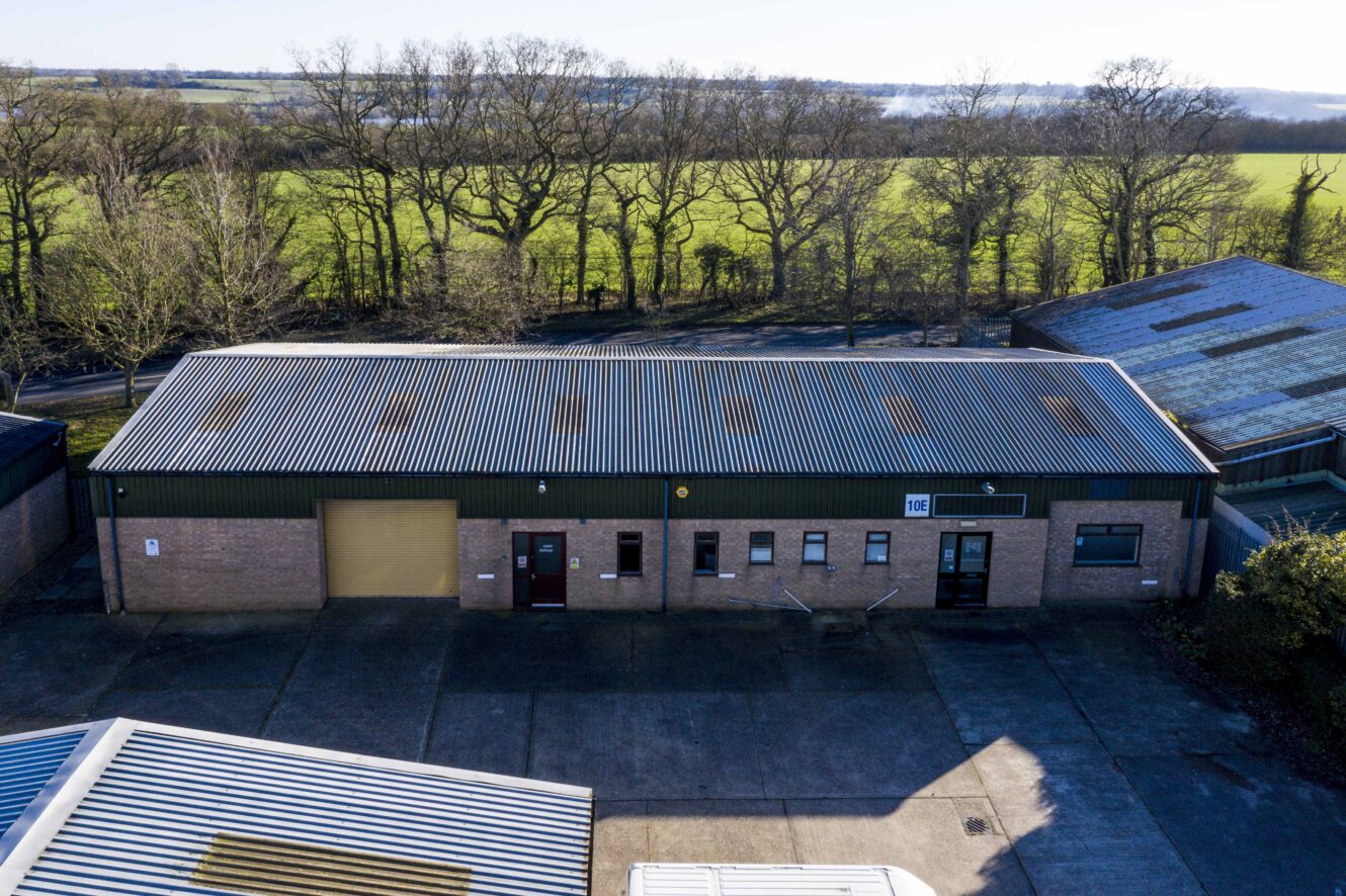 A single-story industrial building with a corrugated metal roof, brick exterior, multiple doors, and a large roller shutter, set near fields and trees.