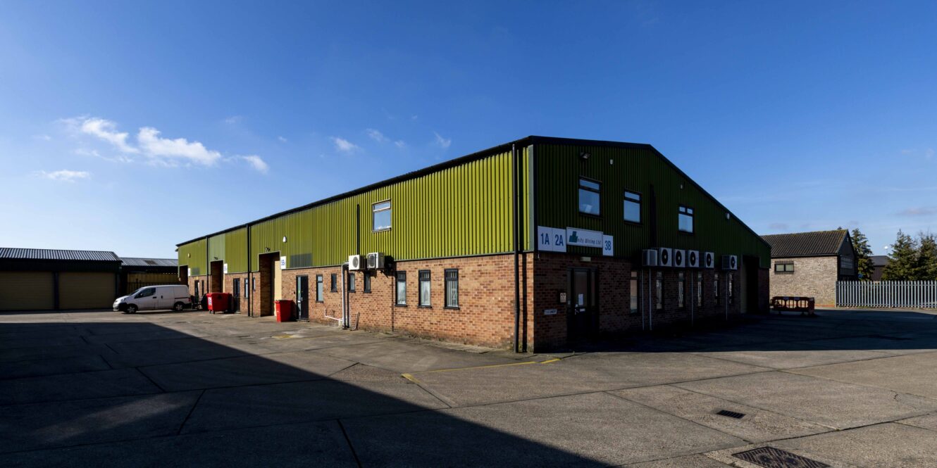A large industrial warehouse with green metal siding and brick lower walls, featuring multiple windows and doors, set in a paved lot under a clear blue sky.