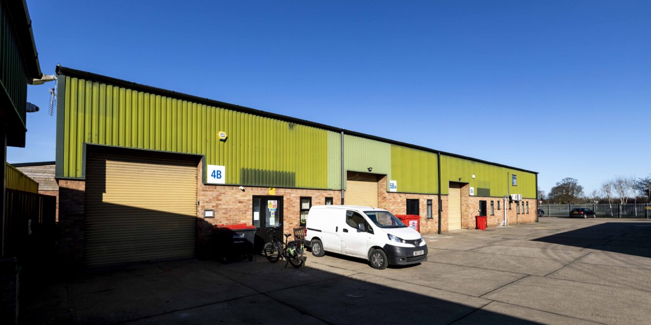 A row of industrial warehouse units with green metal siding, brick walls, and roller doors; a white van and bicycles are parked outside under a clear blue sky.