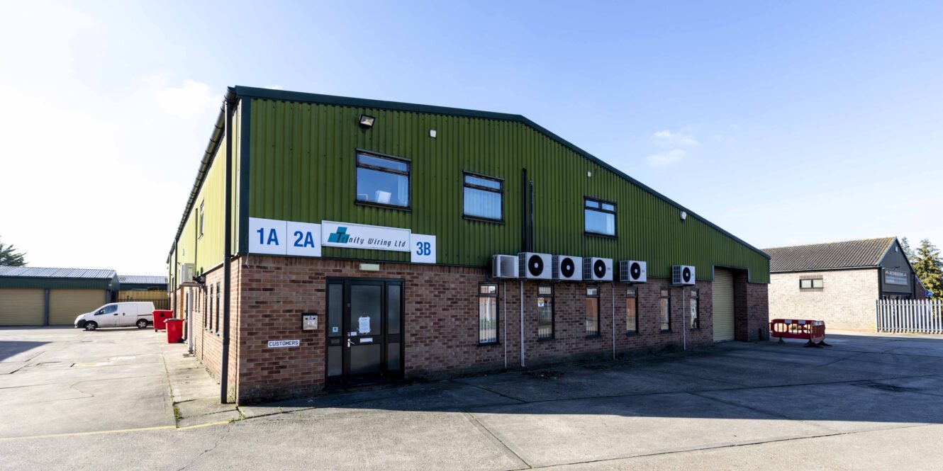 A green and brick industrial building with units labeled 1A, 2A, and 3B; several windows, air conditioning units, and a main entrance at the corner.