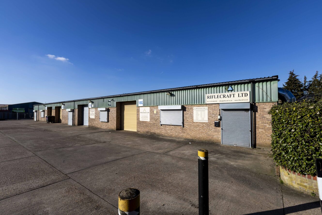 A row of industrial units with brick walls and green metal roofs, including one labeled Riflecraft Ltd, stands on a large, empty concrete lot under a clear blue sky.