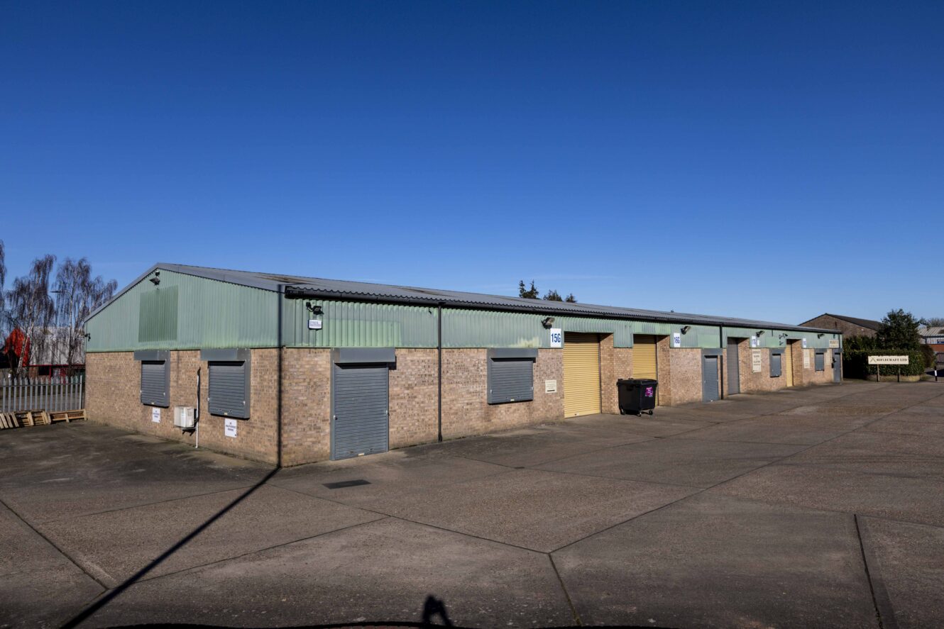 A row of industrial warehouse units with closed roller doors and brick walls under a clear blue sky.