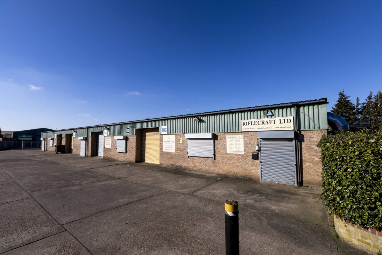A row of industrial units with closed shutters and a sign reading Riflecraft Ltd under a clear blue sky.