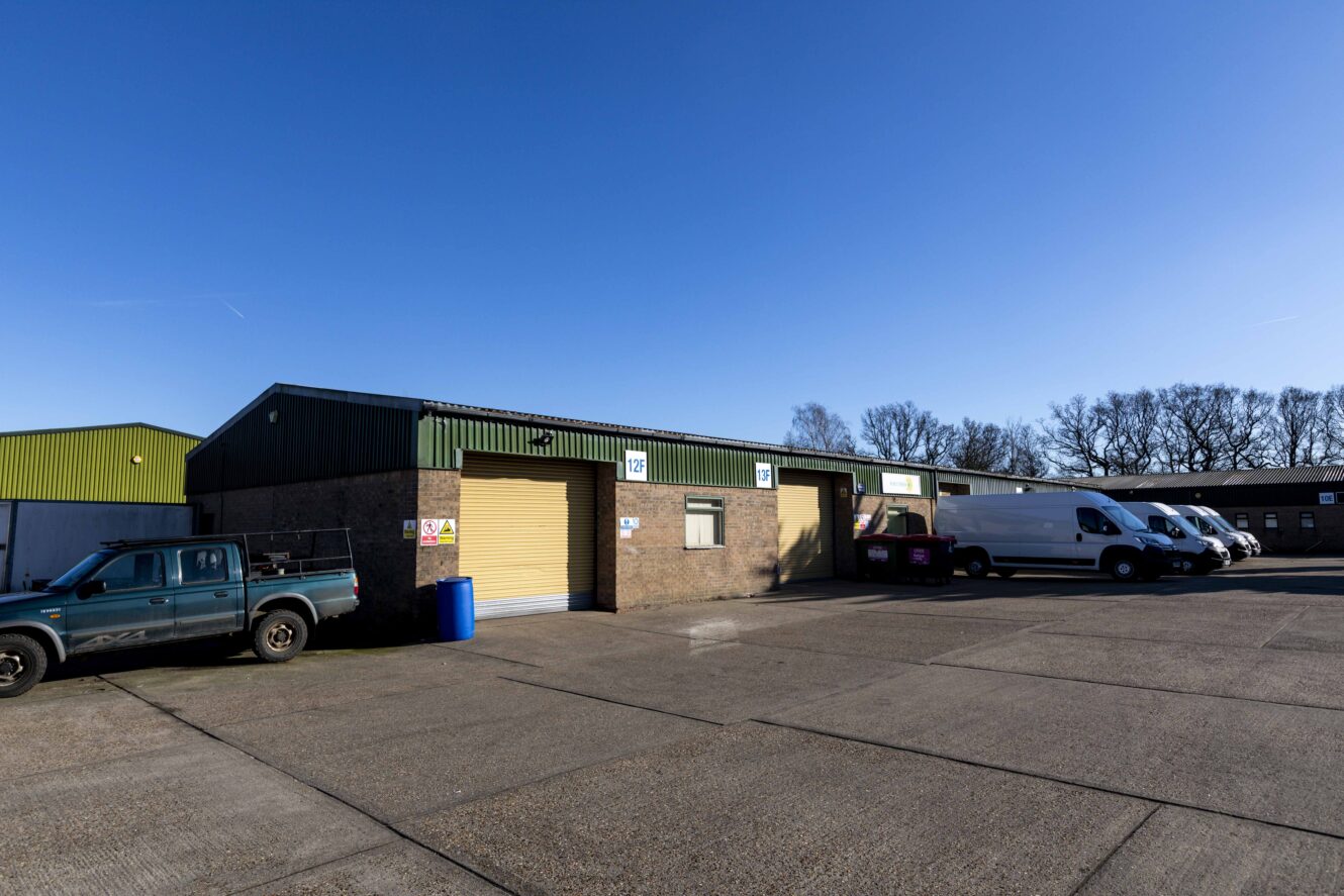 Single-story industrial warehouse with a yellow roller shutter, several parked vehicles, and clear blue sky in the background.