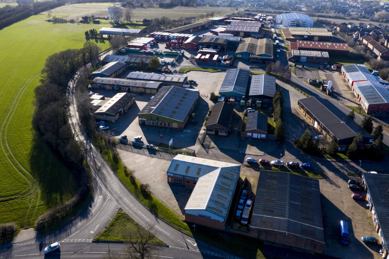 Aerial view of an industrial estate with warehouses, roads, parked vehicles, and surrounding green fields under clear skies.