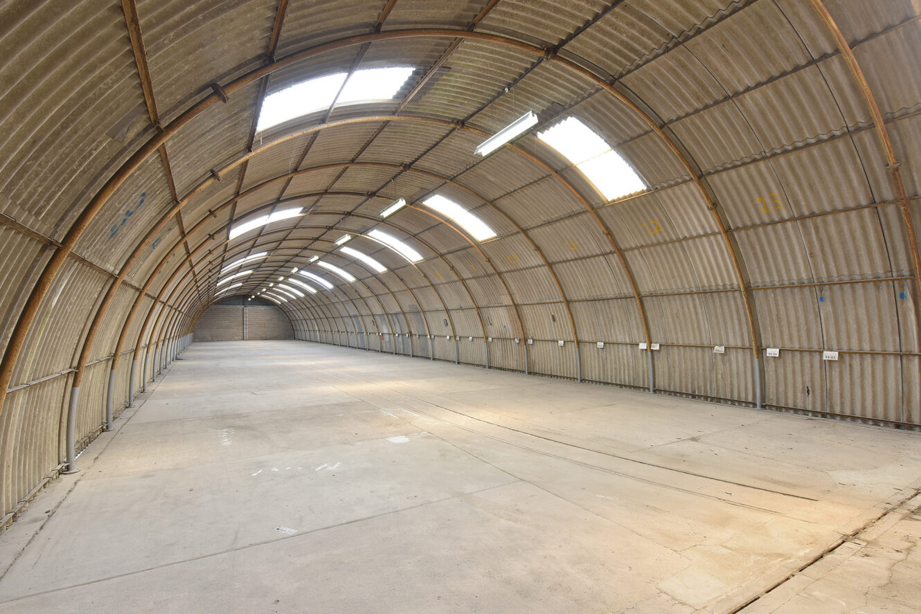 Large, empty, curved-roof warehouse with concrete floor, corrugated metal walls and ceiling, and skylights letting in natural light.