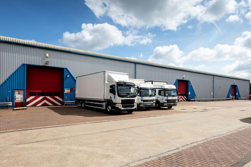Three white delivery trucks are parked in front of red and blue loading bays at a large industrial warehouse on a partly cloudy day.