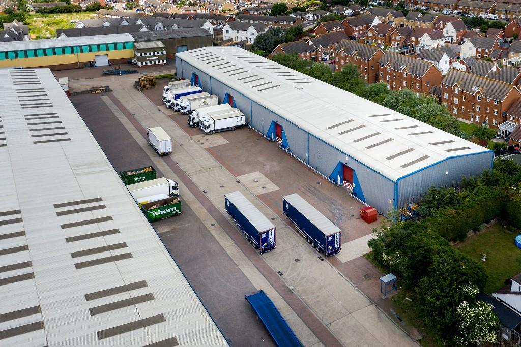 Aerial view of warehouses with trucks and trailers parked outside, surrounded by residential houses and green areas.