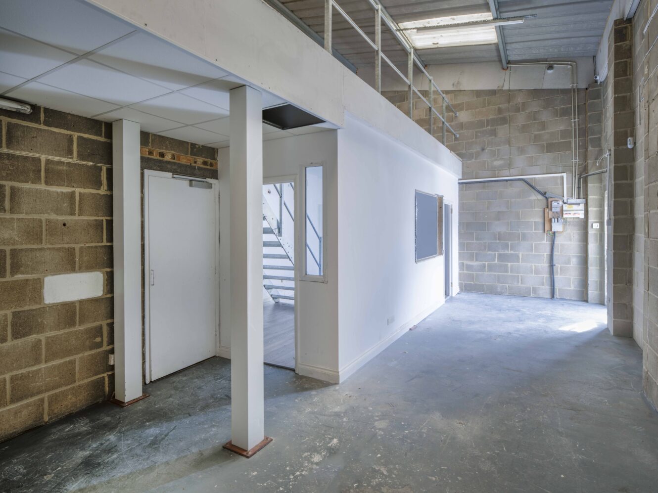 Interior of an empty industrial space with exposed brick walls, concrete floor, a white partition, and a staircase leading to a mezzanine.