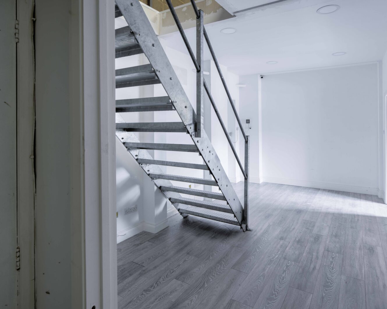 A modern, empty room with light gray wood flooring and a metal staircase leading to an upper level. White walls and ceiling lights are visible.