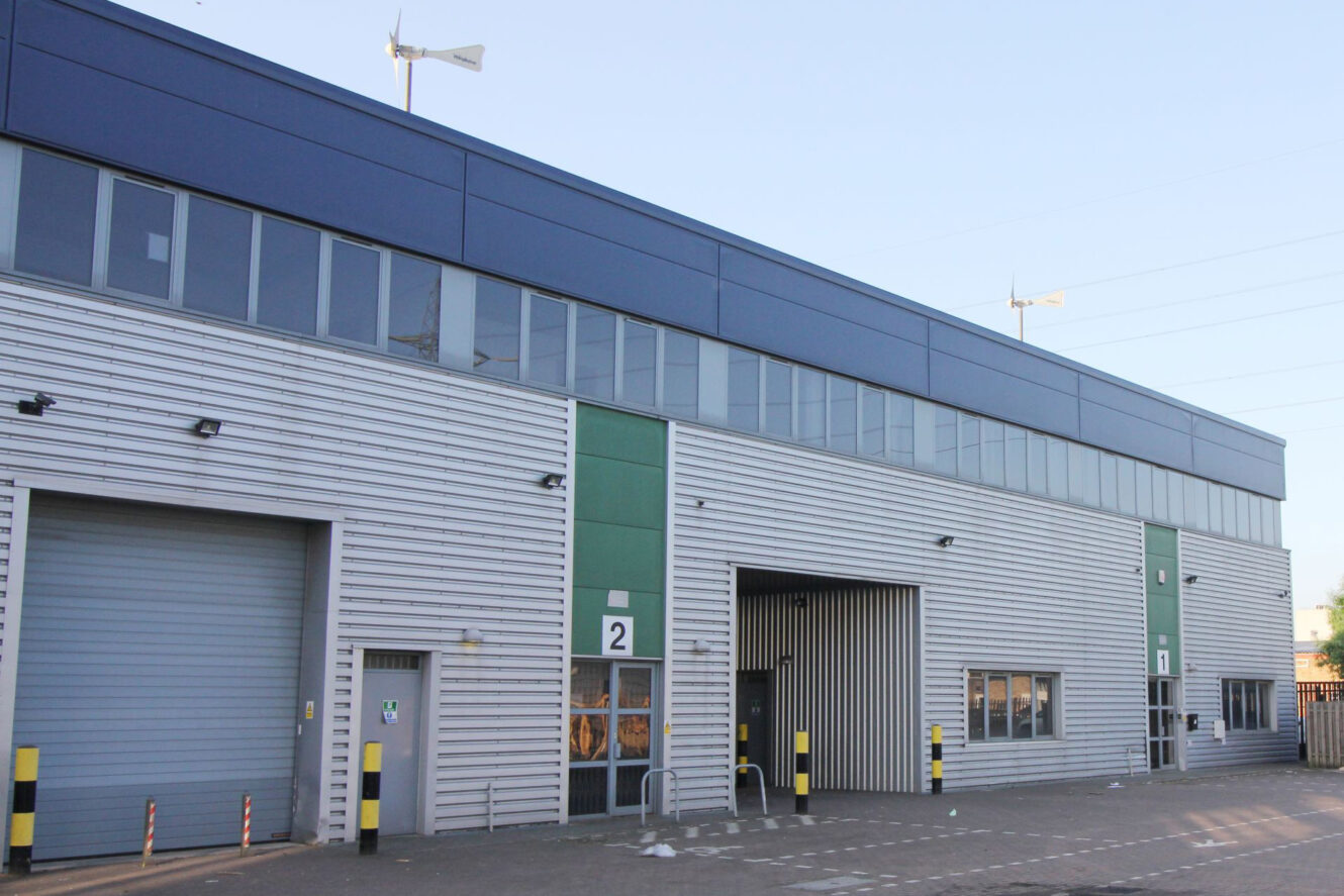 Modern industrial building with metal siding, large windows, two loading bay doors, and numbered entrances labeled 1 and 2. Wind turbines are visible on the roof.
