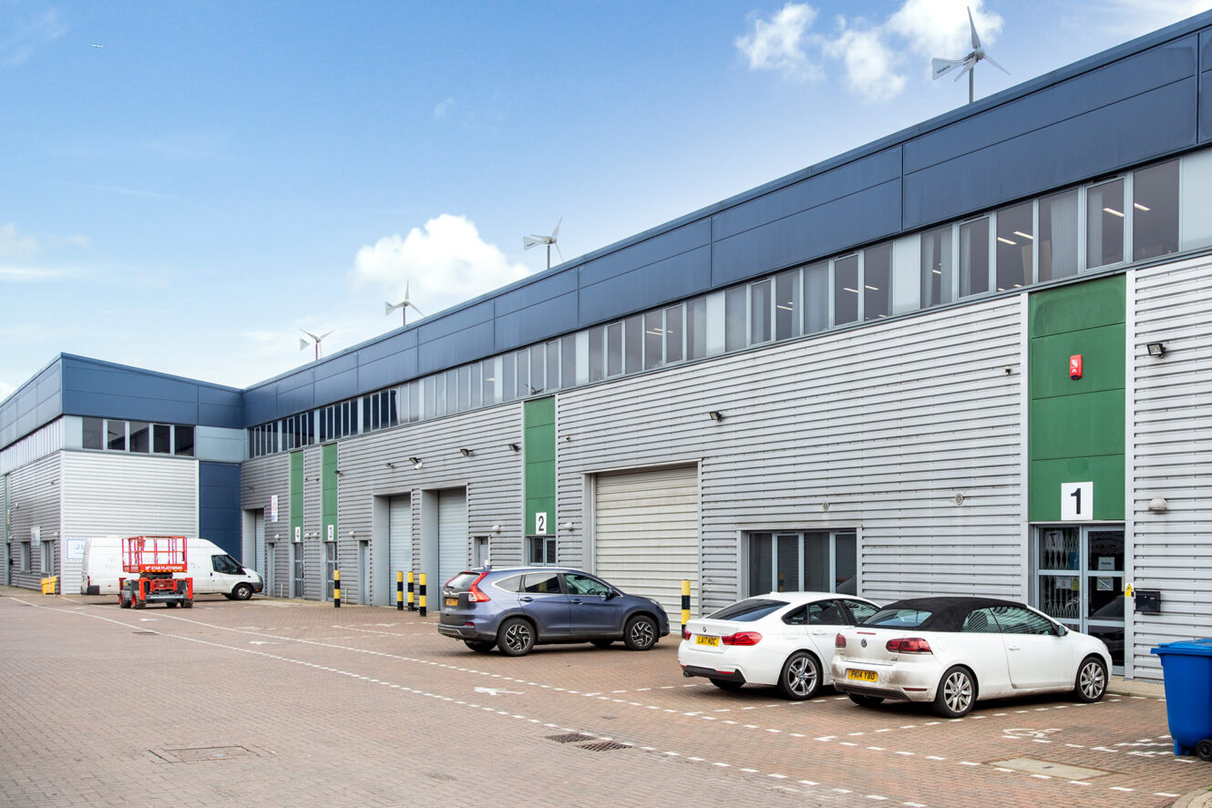 A modern industrial warehouse complex with parked cars, delivery van, and wind turbines on the roof under a blue sky.