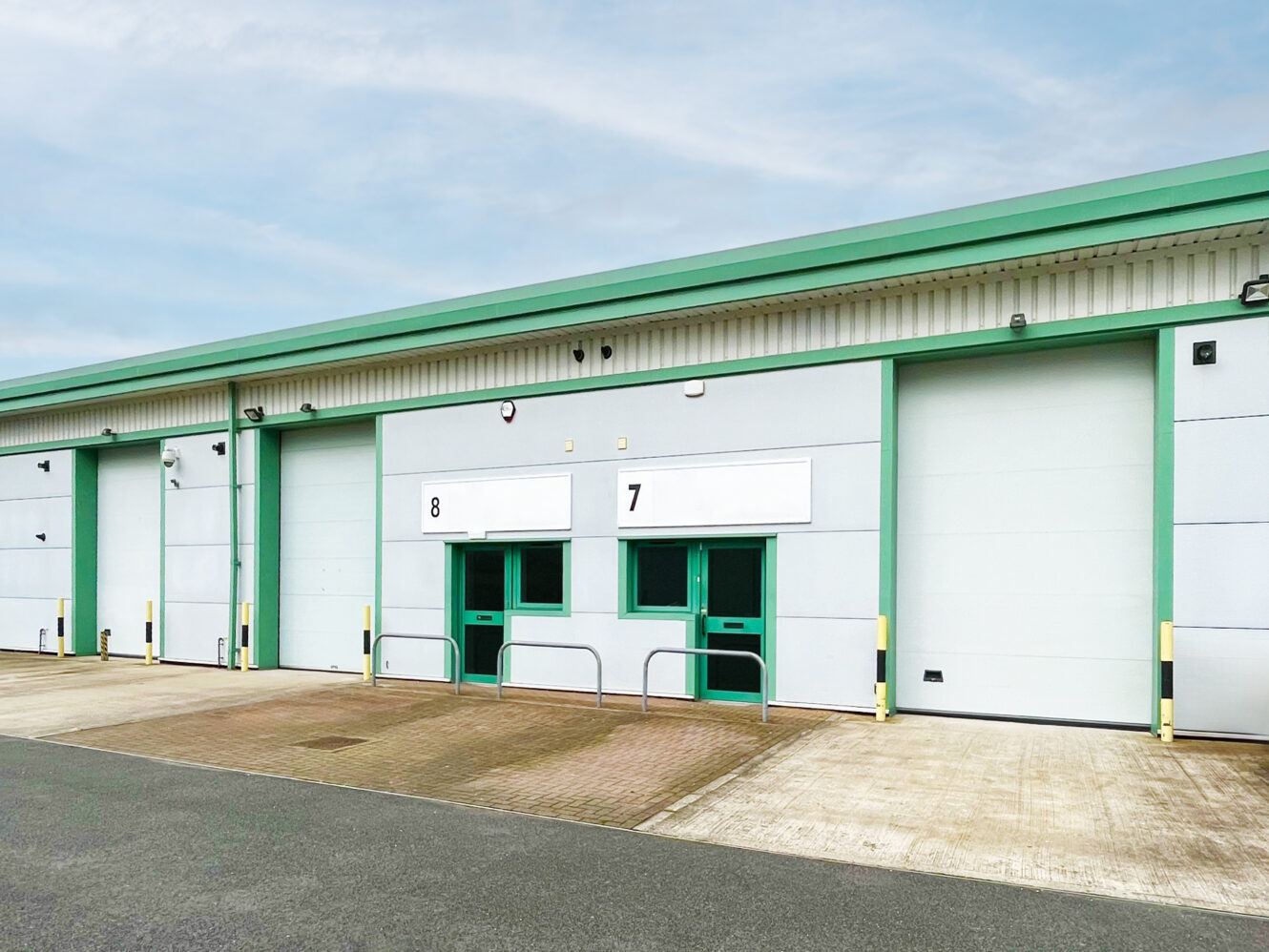 Exterior view of an industrial warehouse with green trim, featuring large closed roller doors labeled 7 and 8, and a paved area in front.