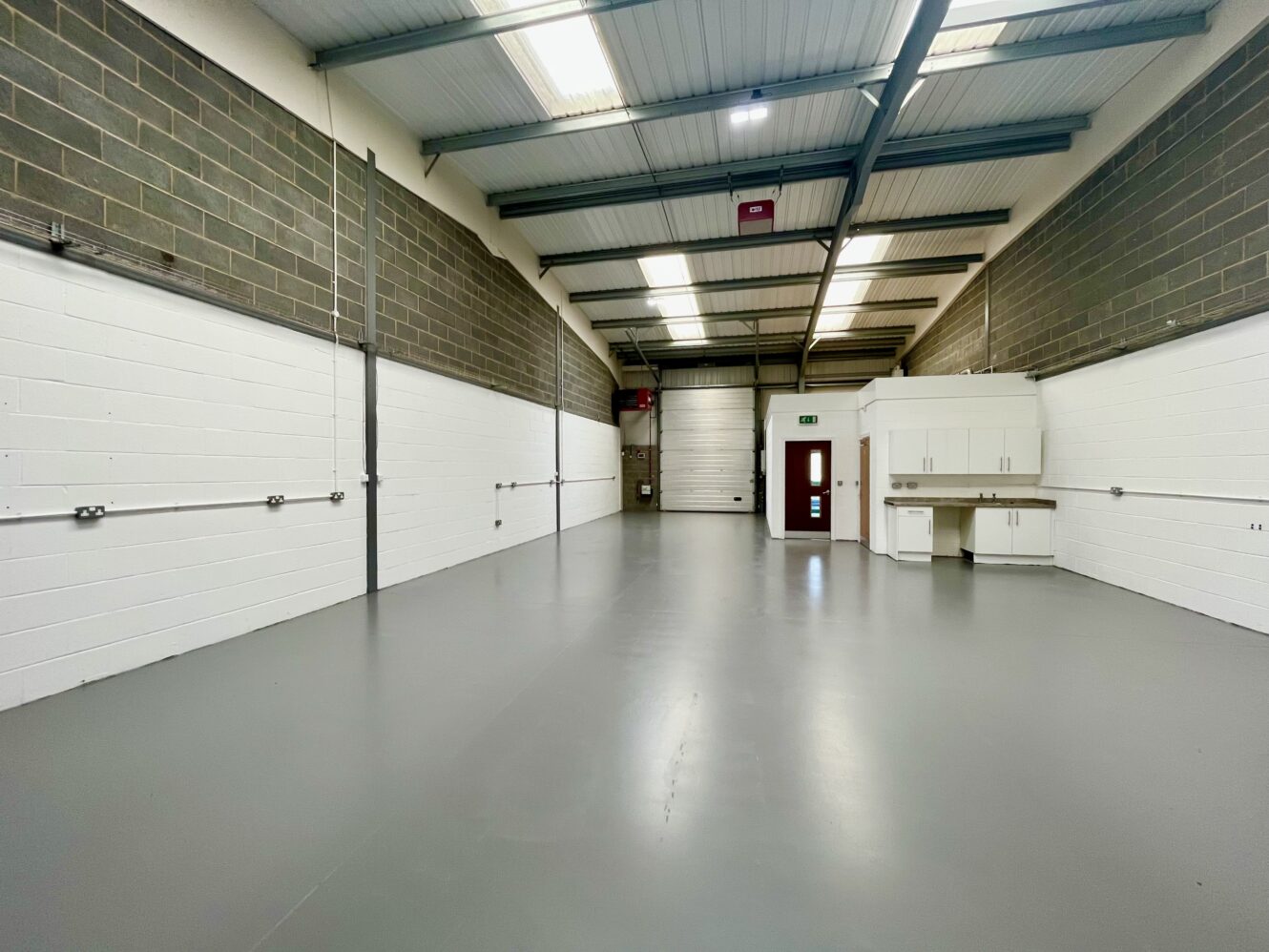 Interior of an empty industrial warehouse with white walls, grey floor, high ceiling, overhead lights, a red door, and storage cabinets on the right.