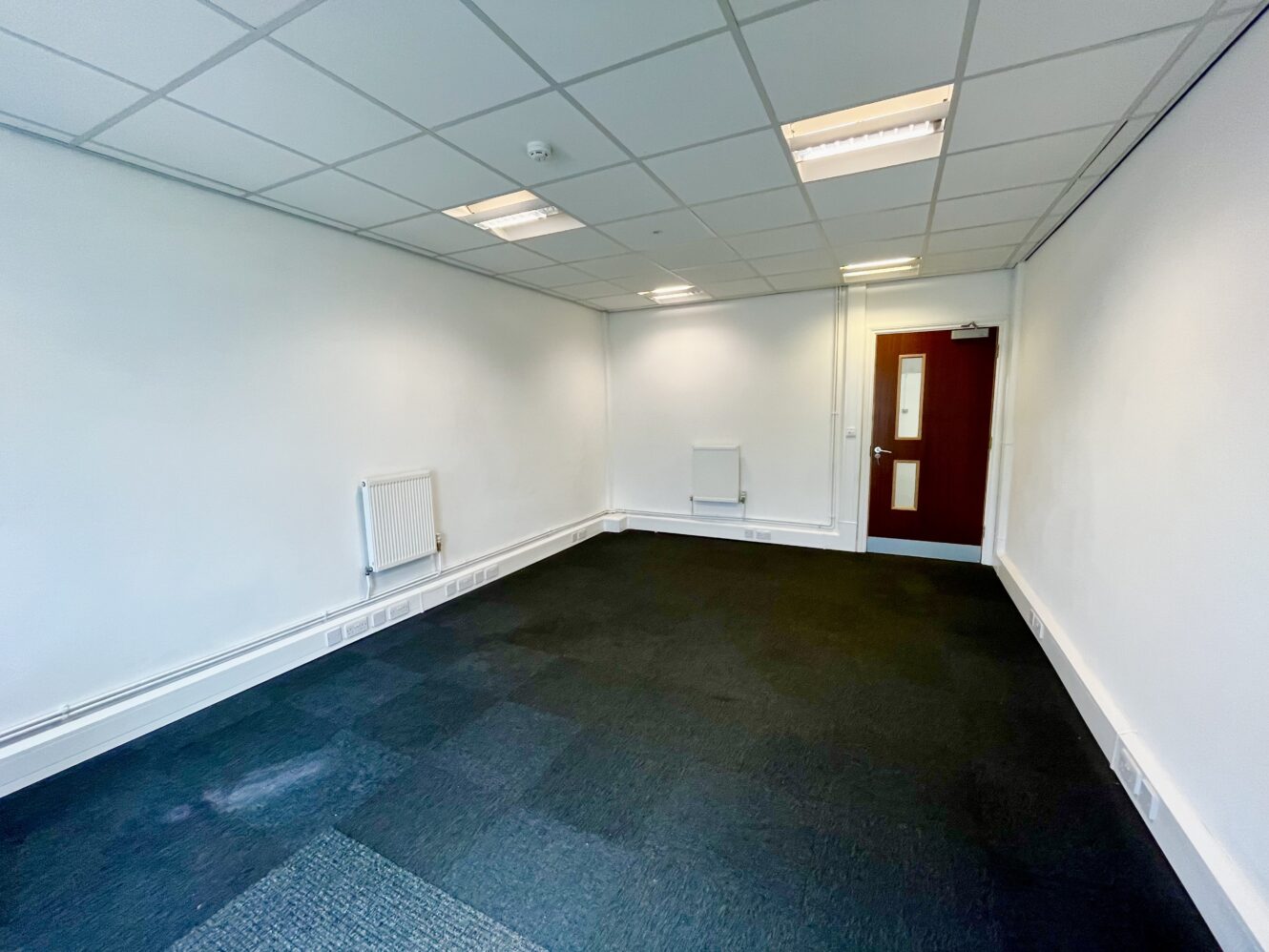 Empty office room with white walls, black carpet flooring, a red door with window, two radiators, ceiling lights, and a drop ceiling.
