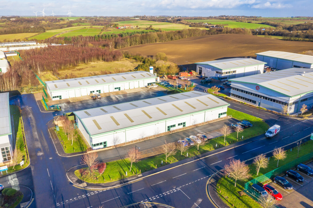 Aerial view of an industrial park with several large warehouse buildings, parking lots, nearby roads, and green fields in the background.