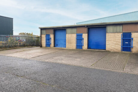 Industrial building with beige brick walls, three blue roller shutter doors, and matching blue entry doors, located next to a paved road and fenced area.