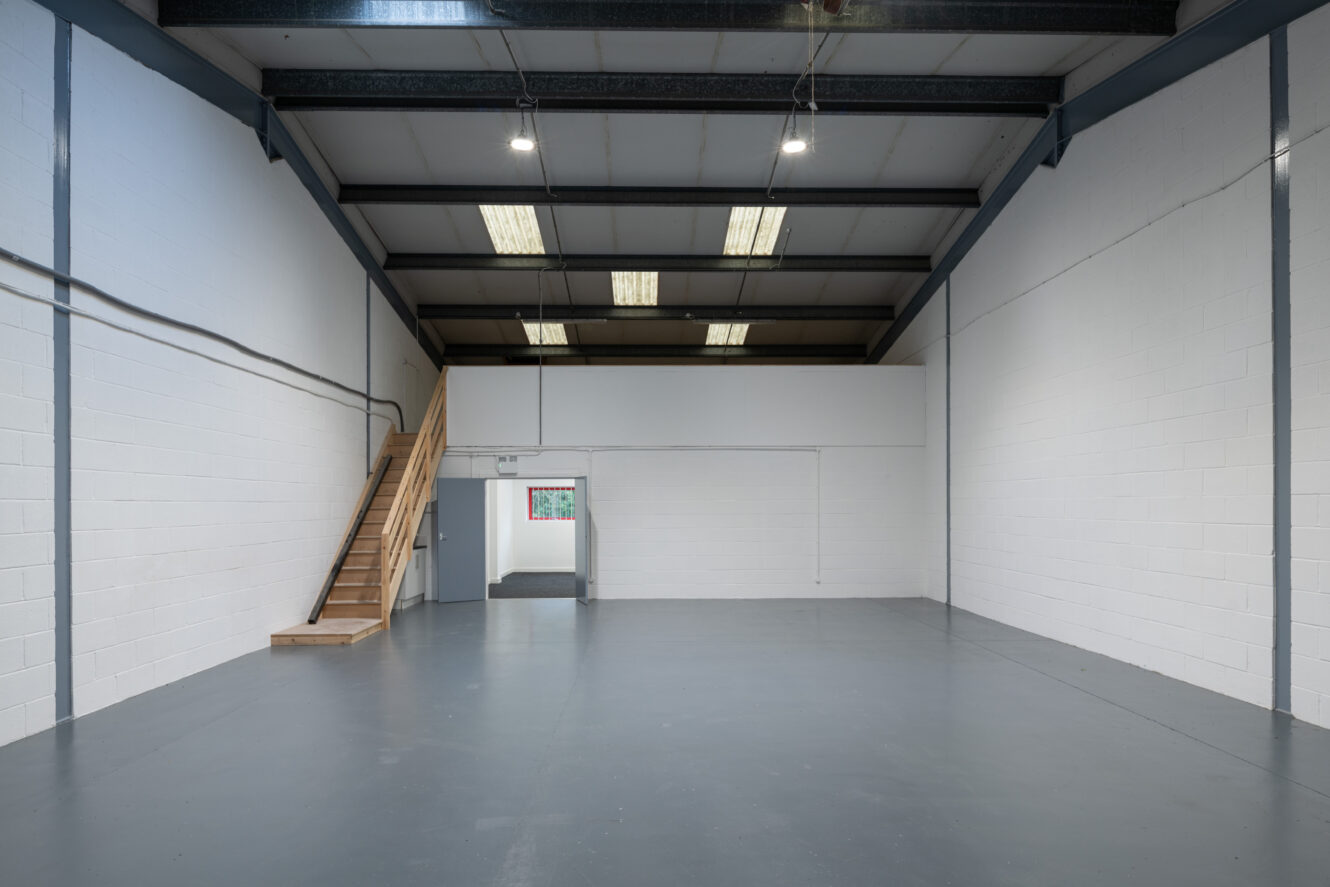Empty industrial warehouse unit with white painted brick walls, grey concrete floor, exposed ceiling beams, fluorescent lights, and a wooden staircase leading to a mezzanine level.