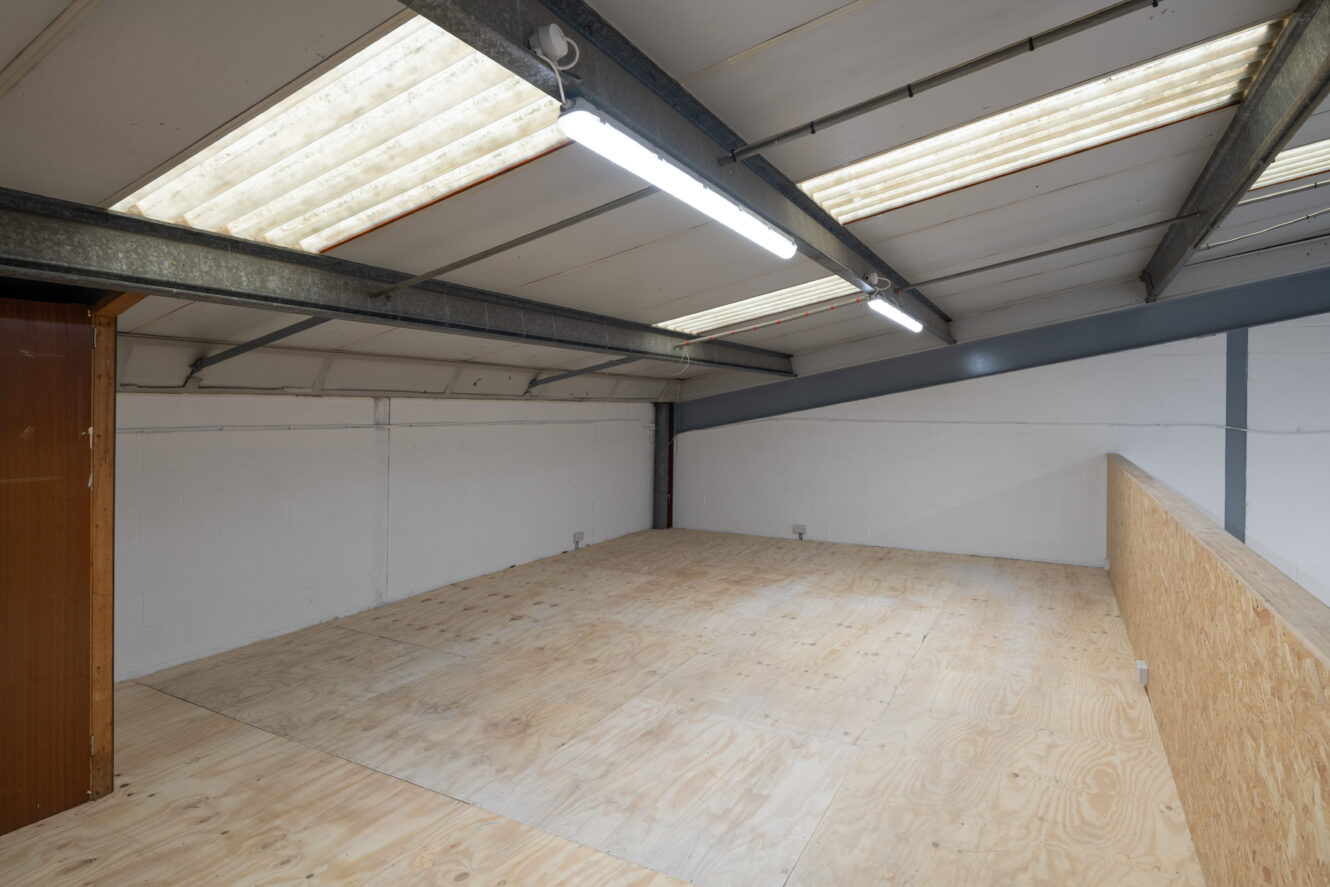 Empty industrial mezzanine with plywood flooring, white painted walls, exposed metal beams, fluorescent lighting, and skylight roof panels.