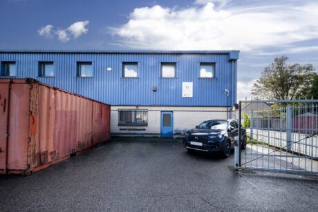 A blue industrial building with several small windows, a red shipping container, a black car parked by the entrance, and a metal gate partially open.