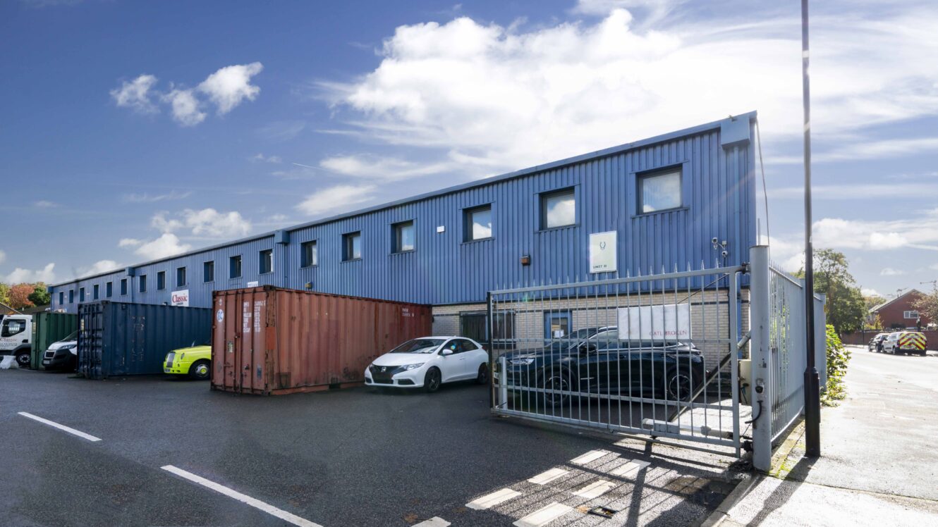 A blue industrial warehouse with parked cars and shipping containers outside, viewed from behind a metal gate under a partly cloudy sky.