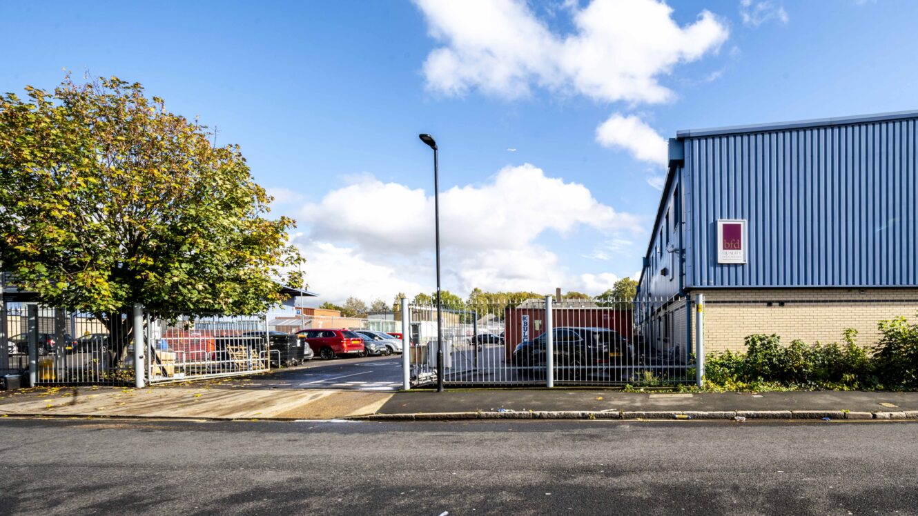 Industrial estate entrance with metal gates, a blue warehouse building on the right, parked vehicles, and a tree on the left under a partly cloudy sky.