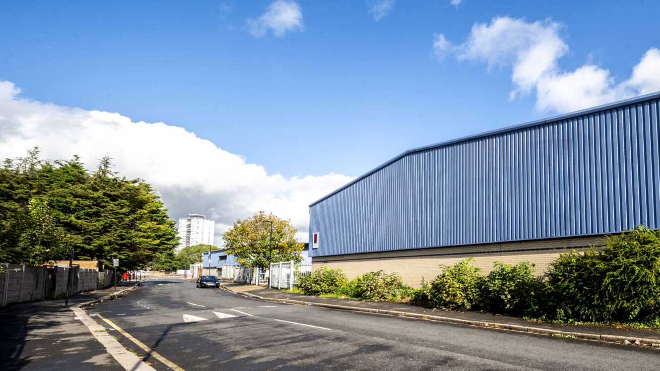 A quiet street runs past a large blue industrial warehouse with trees and bushes lining the sidewalk under a partly cloudy sky.