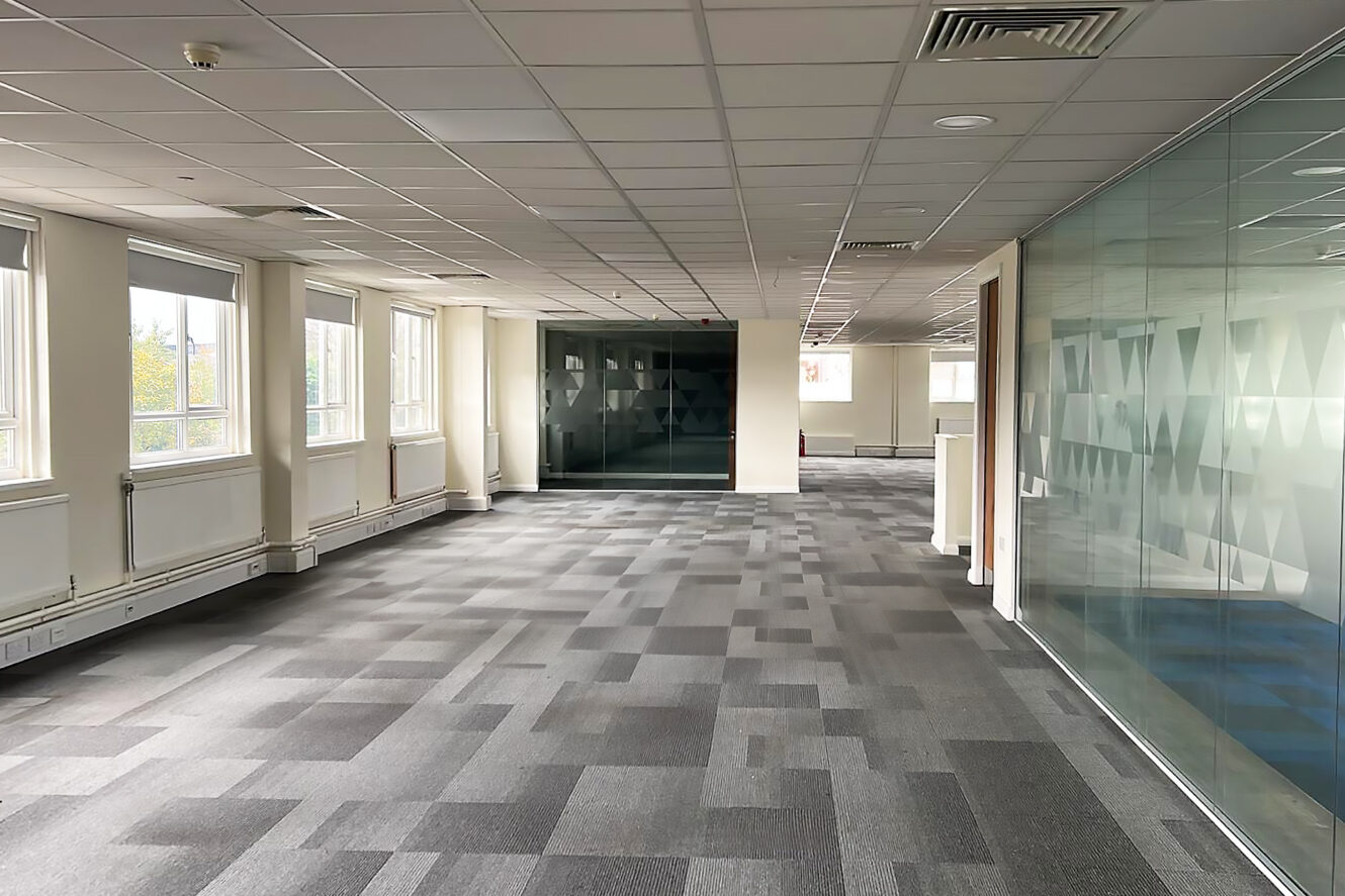An empty modern office space with large windows, gray carpeted floor, white walls, and a glass partition on the right side.