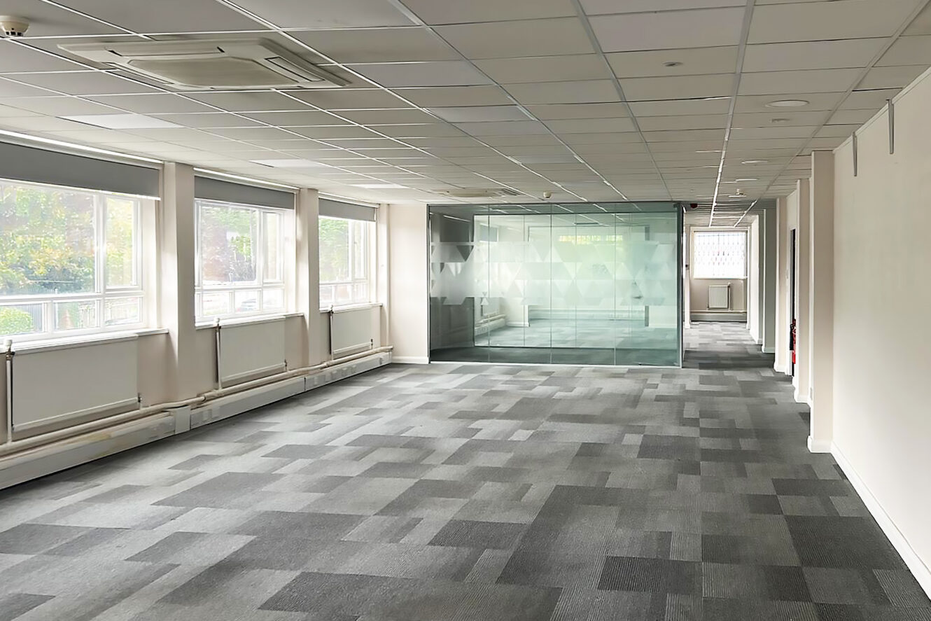 Empty office space with gray carpet tiles, large windows on the left, and a glass-walled meeting room at the far end.