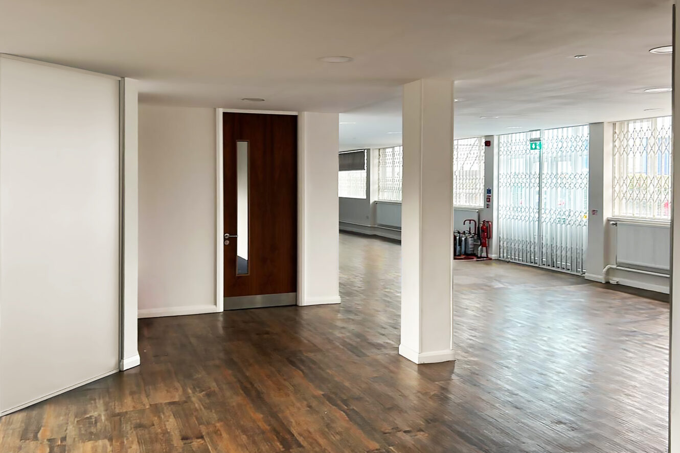Empty office space with wooden flooring, white walls, a brown door, large windows with security bars, and a fire extinguisher near an emergency exit.
